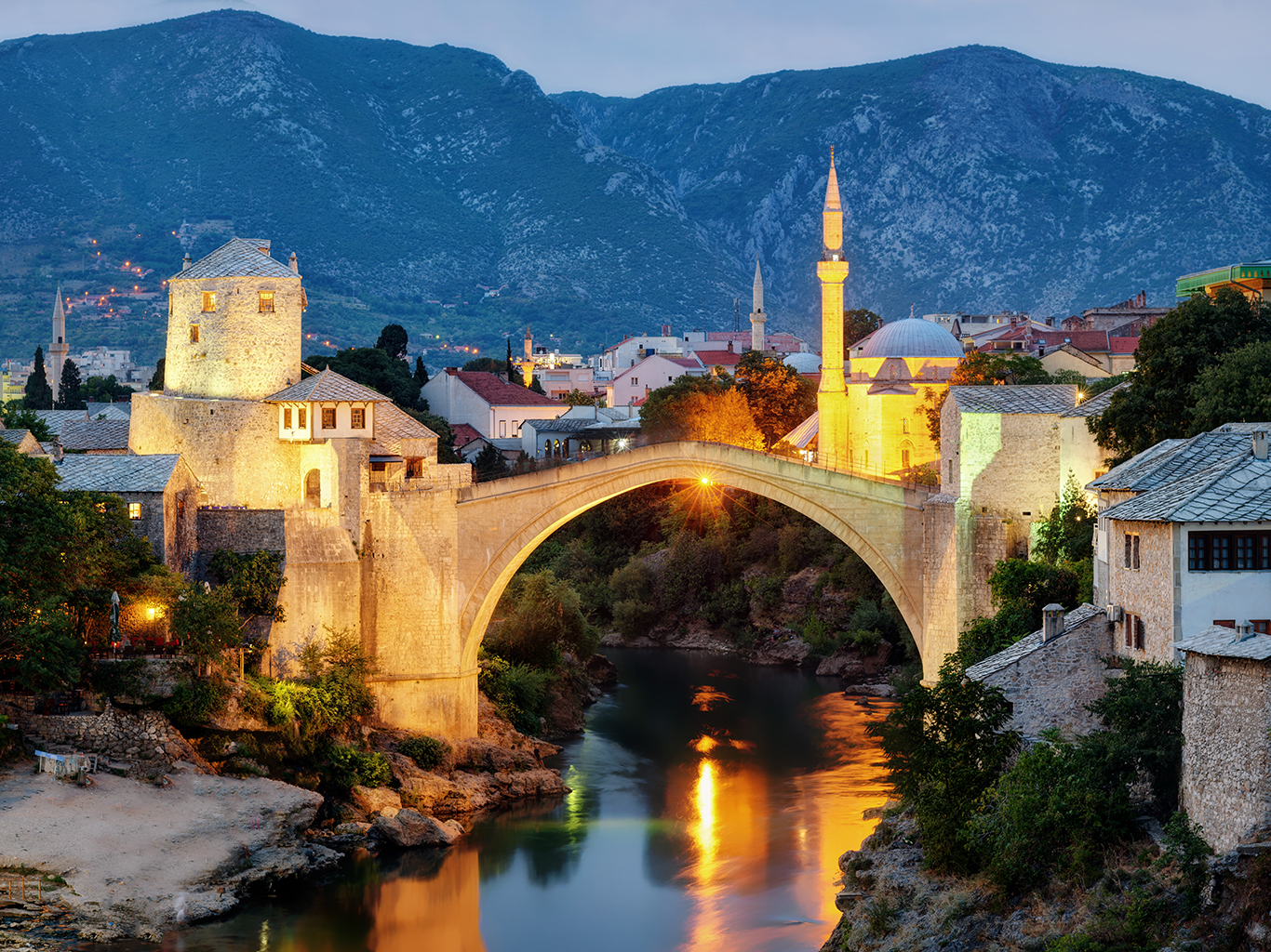 Večerní Mostar s osvětleným mostem, strážní věží a minaretem v pozadí