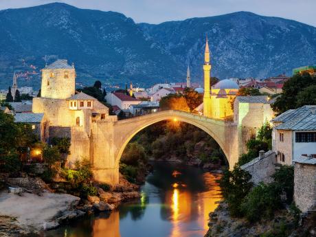Večerní Mostar s osvětleným mostem, strážní věží a minaretem v pozadí